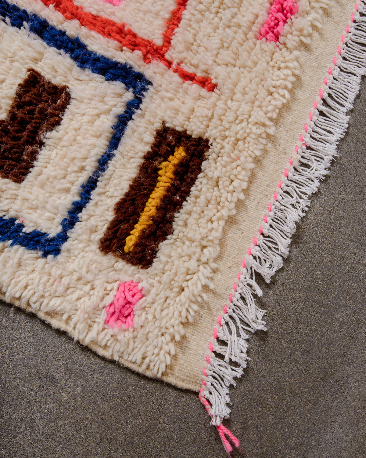 Corner of Azilal rug with white cotton tassels laying on concrete floor.